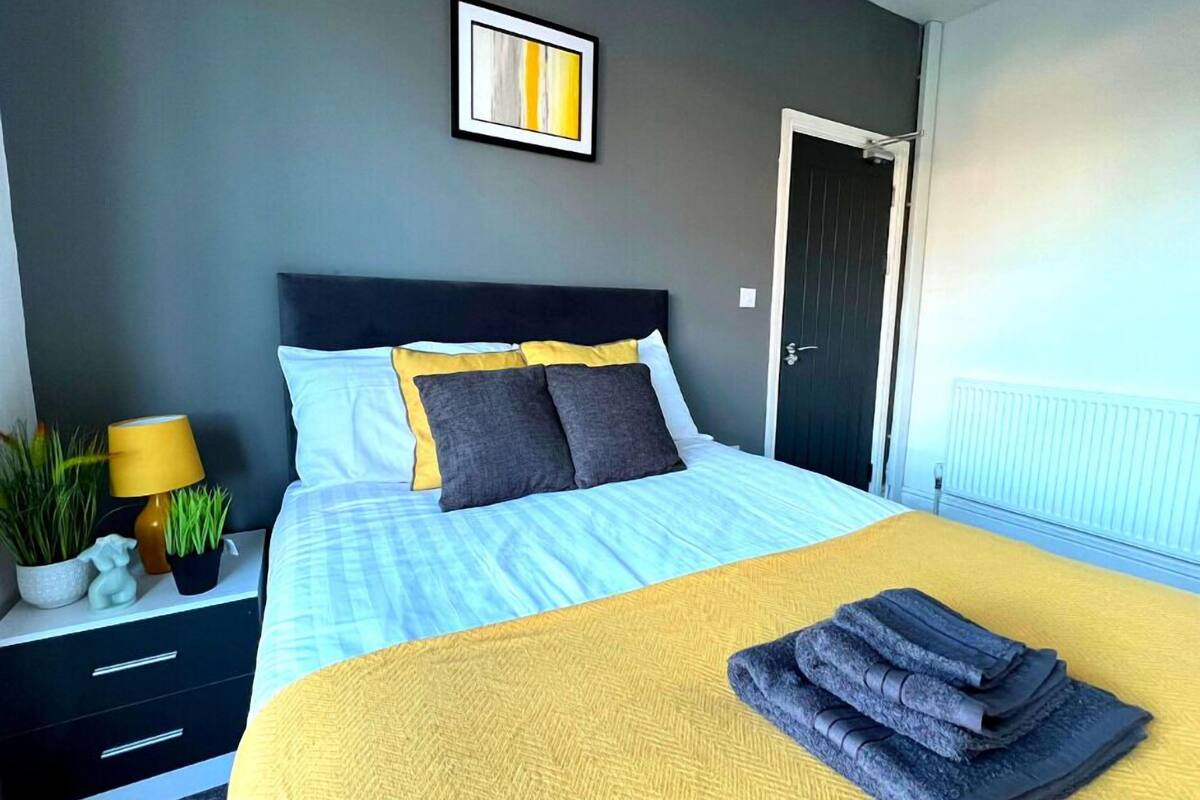 Un lit bien fait, avec une couverture jaune et des serviettes pliées, une table de chevet avec une lampe et une plante en pot, et un tableau encadré accroché au mur.