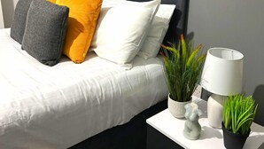 A neatly made bed with white and gray bedding, a black headboard, and various pillows. There are two potted plants on a bedside table, a white lamp, and a small decorative item. - Chantry Suites by SA Stays (Grimsby)