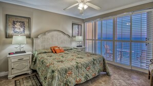 Un dormitorio con cama, mesitas de noche, ventilador de techo, ventana con persianas y vistas al exterior.
