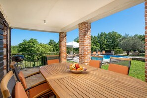 An outdoor patio with a wooden table, orange chairs, a grill, and a swimming pool in the background.