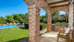 A brick archway leading to a pool area with a grassy lawn and a wooden chair.