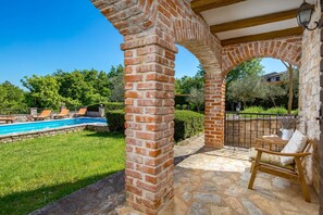 Un arco de ladrillo que da acceso a una zona de piscina con césped y una silla de madera.