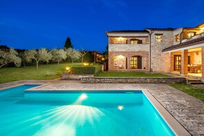 Una casa de dos pisos con piscina, fachada de piedra e iluminación exterior.