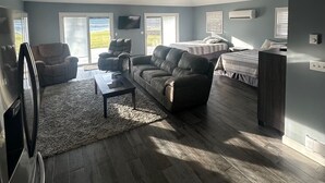 Ein geräumiges Wohnzimmer mit einer großen Couch, einem Couchtisch und einem Fernseher. Es gibt zwei Fenster mit Jalousien, einen Deckenventilator und einen Kamin.