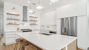 A modern kitchen with white cabinetry, a stainless steel refrigerator, wooden stools, and pendant lighting. - New Luxury Beach Home with Private Pool (Panama City Beach)