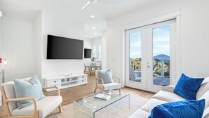 A modern living room with a flat-screen TV, a glass coffee table, and a sofa with blue cushions. - New Luxury Beach Home with Private Pool (Panama City Beach)