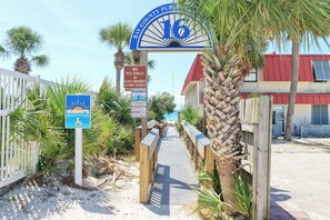 Ein Schild, das den Zugang zum Strand kennzeichnet, mit einem Rettungsturm, einer Holzpromenade, die zum Strand führt, und einem roten Gebäude im Hintergrund.
