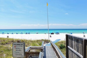 Ein Strand mit einer Holzpromenade, die zum Ufer führt, bewölkter Himmel und ein Schild mit Hinweisen zu Strand-Warnflaggen.