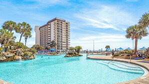 A large outdoor swimming pool with a clear blue water, surrounded by palm trees and a hotel building in the background. - Tropical Beachfront Retreat (Panama City Beach)