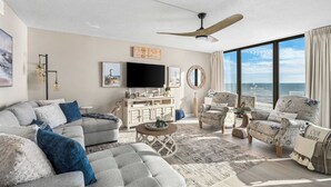 Ein geräumiges Wohnzimmer mit großem Fenster zum Meer, Deckenventilator und Fernseher.