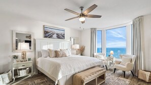 Un dormitorio con una cama grande, ventilador de techo, vista al océano y una ventana con cortinas.