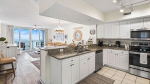 Una cocina moderna con armarios blancos, encimeras de granito y electrodomésticos de acero inoxidable. Hay una isla grande con un fregadero, y la cocina se abre a un balcón con vista al océano.