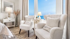 Un dormitorio moderno con dos sillones blancos, una mesita de noche con lámpara, un espejo y vistas al mar.