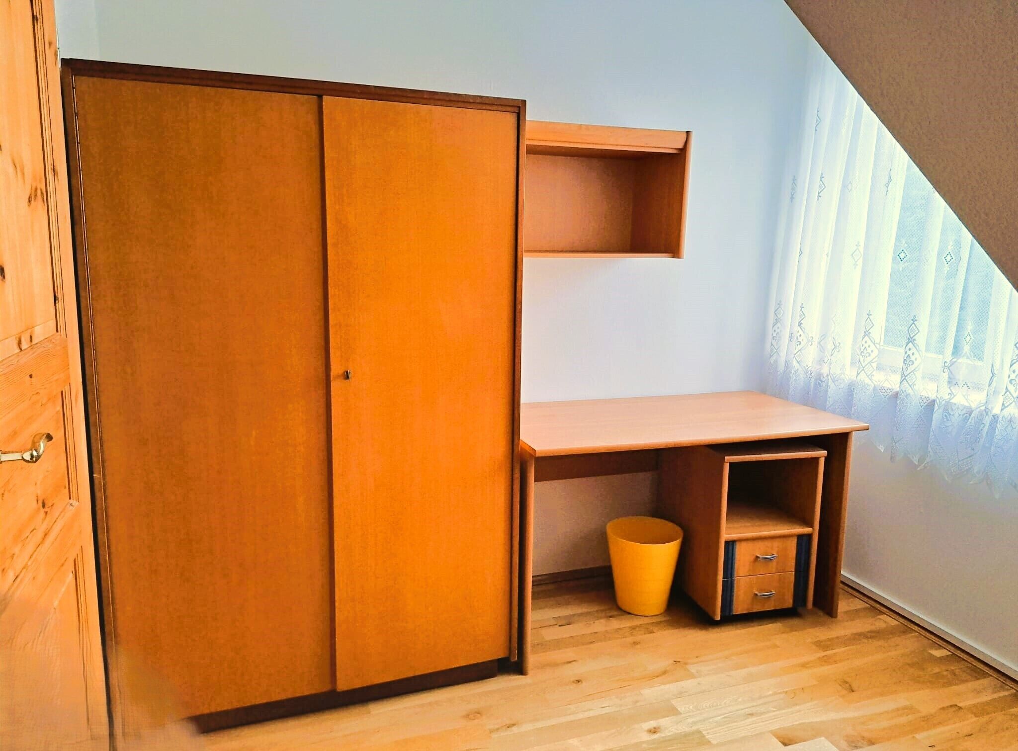 Ein Zimmer mit Holzschrank, Schreibtisch mit Schubladen, kleiner Mülleimer und Fenster mit durchsichtigen Vorhängen.
