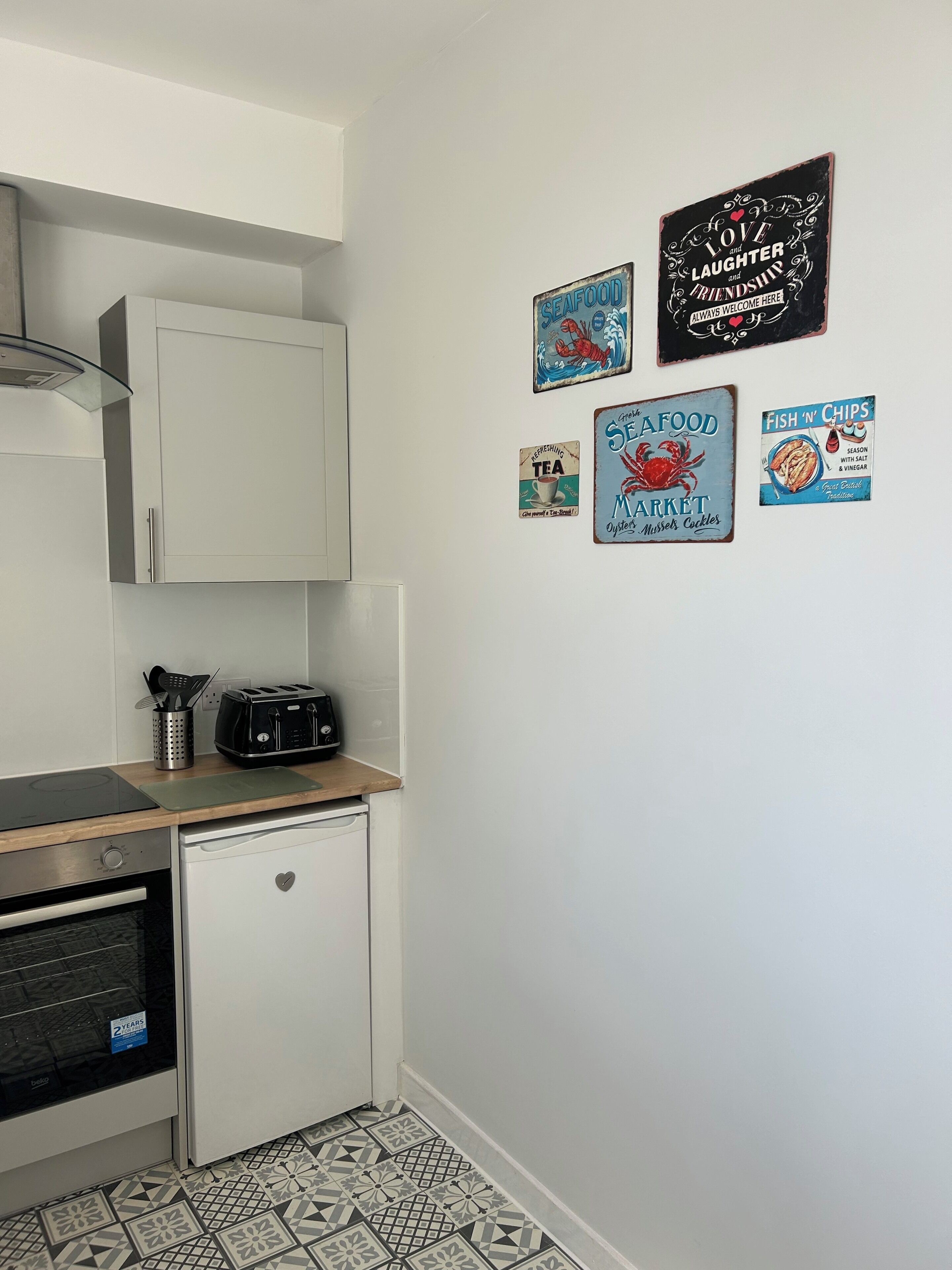 Eine kleine Küche mit weißen Schränken, einem Toaster und einem kleinen Kühlschrank. An den Wänden sind maritime Dekorationen und ein gemusterter Boden zu sehen.