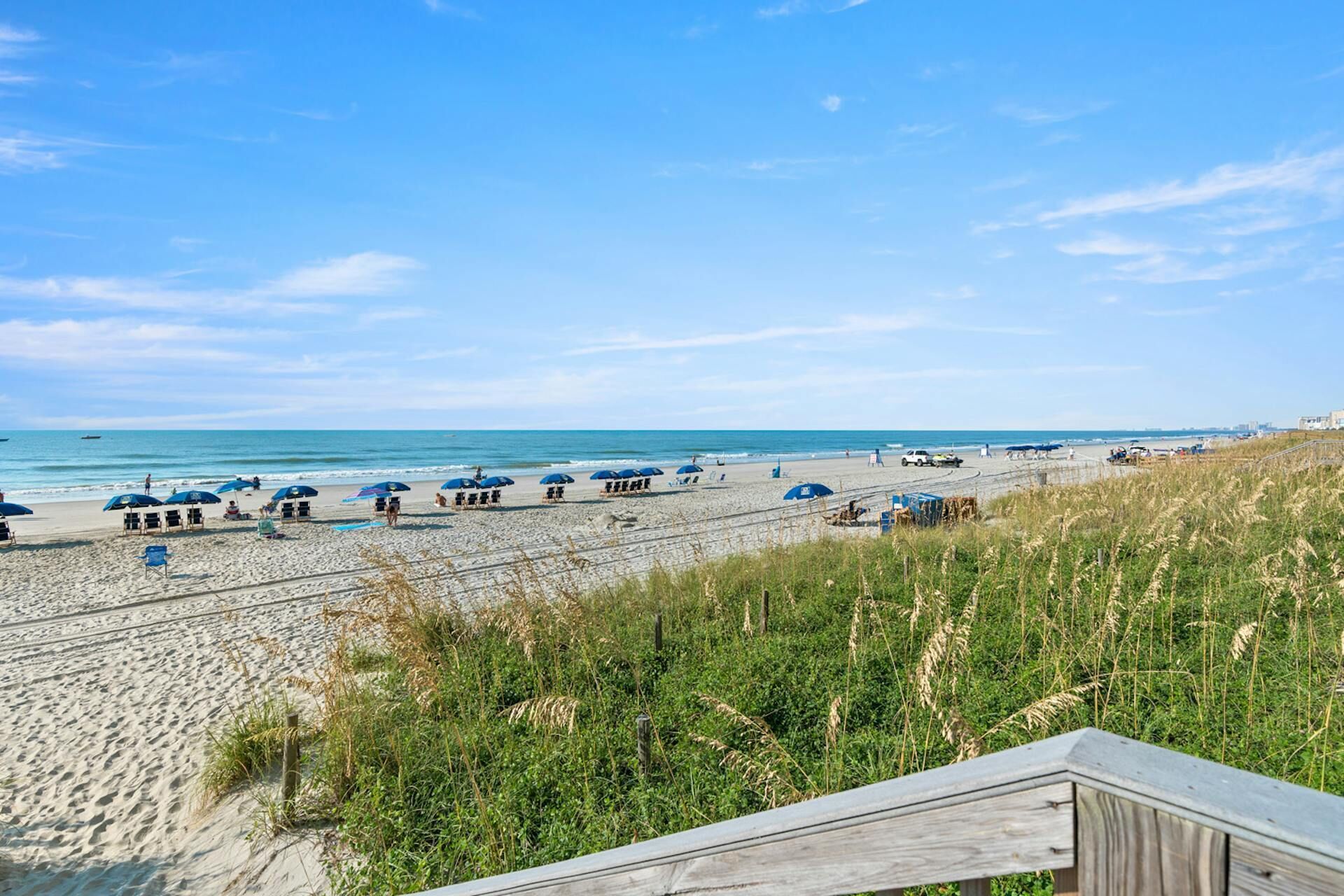 A beach with blue umbrellas, a sandy shore, and a grassy area with a wooden deck.
