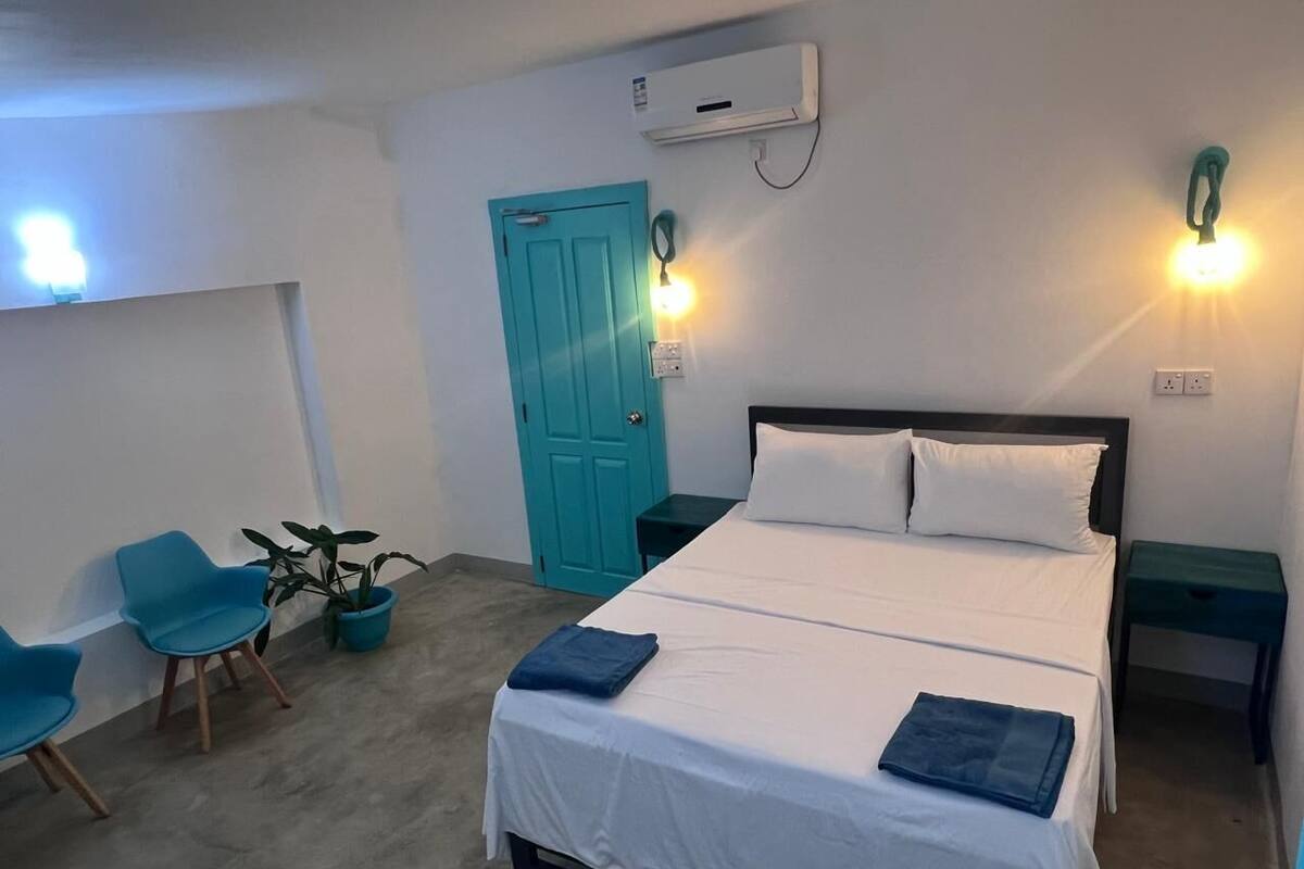 Ein Schlafzimmer mit einem Bett, zwei Nachttischen, einer blauen Tür, einer Topfpflanze und zwei blauen Stühlen.