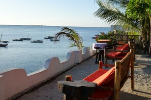 A beachside seating area with wooden chairs and tables, overlooking a calm sea with several boats.