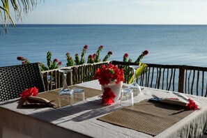 A table set for dining with a view of the sea, a vase of red flowers, and a wicker chair.