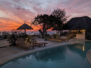 A poolside area with lounge chairs and a thatched-roof structure, overlooking a beach at sunset.