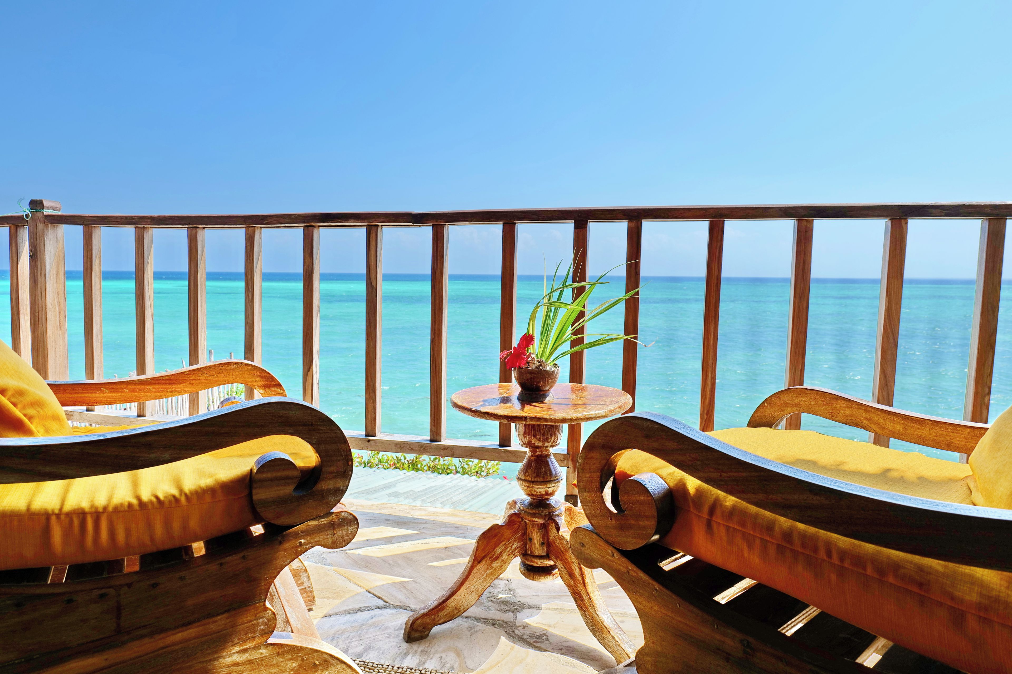 A wooden deck with a view of the ocean, two wooden chairs with yellow cushions, and a small round table with a potted plant.