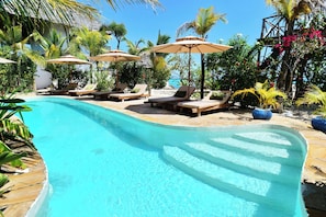 A swimming pool with lounge chairs and umbrellas, surrounded by tropical plants and a clear sky. - Lala Land Lodge (Kizimkazi)