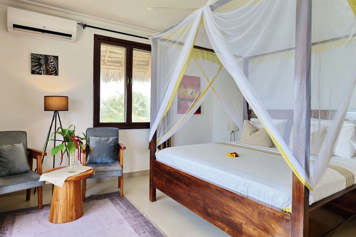 A bedroom with a four-poster bed, a sitting area with a chair and a small table, and a large window.