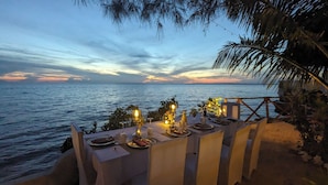 Une table dressée pour un repas sur une plage, avec vue sur la mer et un coucher de soleil en arrière-plan.