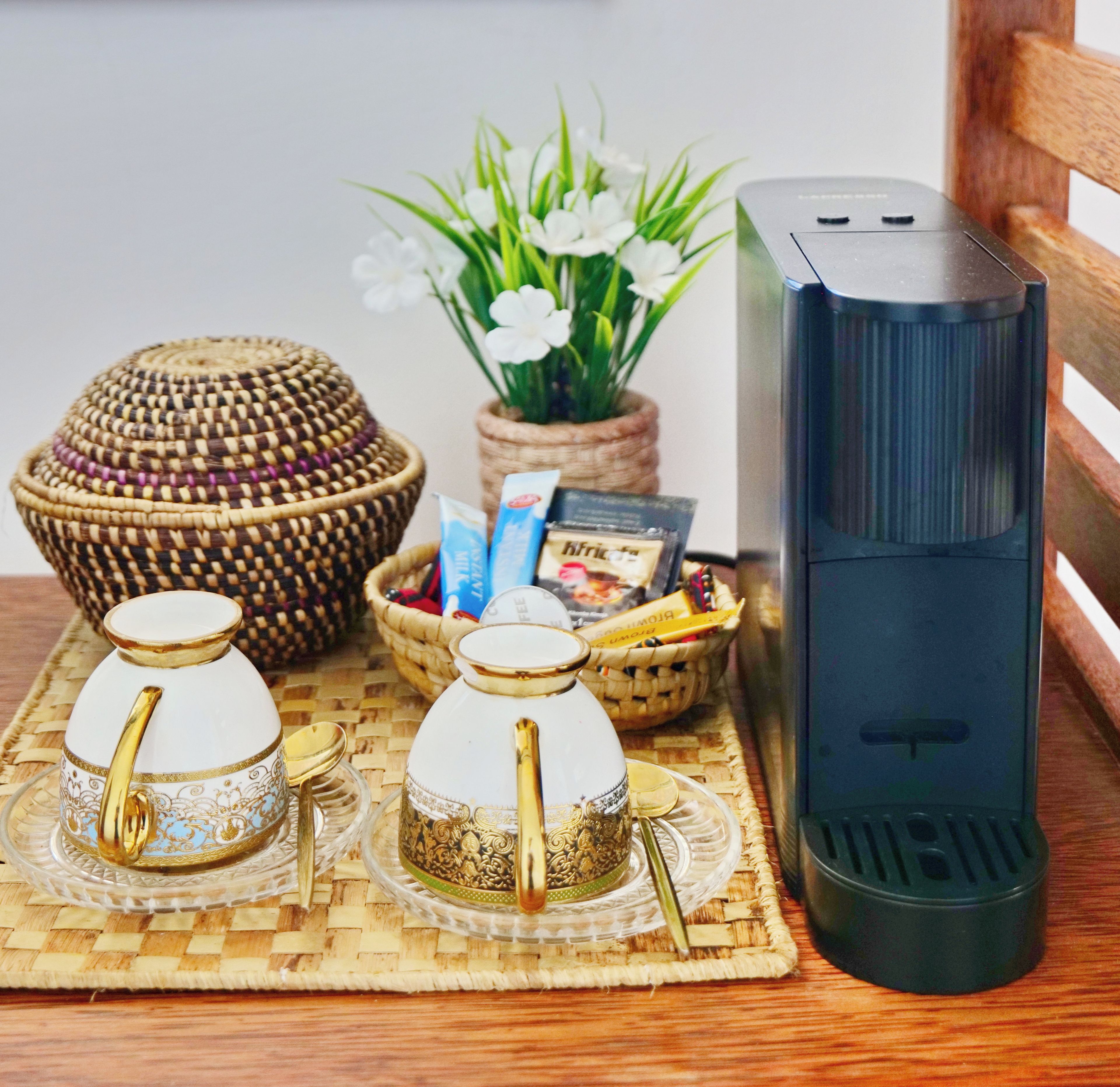 A table with a coffee maker, two teapots, a woven basket, a potted plant, and a box of tea.