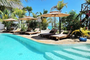 A poolside area with lounge chairs and umbrellas, surrounded by tropical plants and a clear view of the sea. - Lala Land Lodge (Kizimkazi)