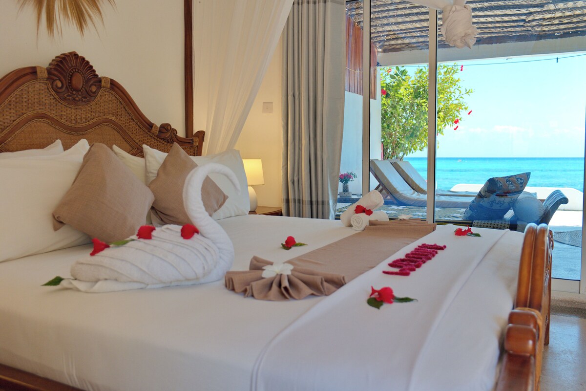 A hotel room with a bed, a view of the sea, and a balcony with lounge chairs.