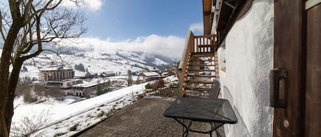 Un área exterior cubierta de nieve con una escalera de madera que conduce a un edificio, un camino pavimentado y una vista de un pueblo nevado en la distancia.