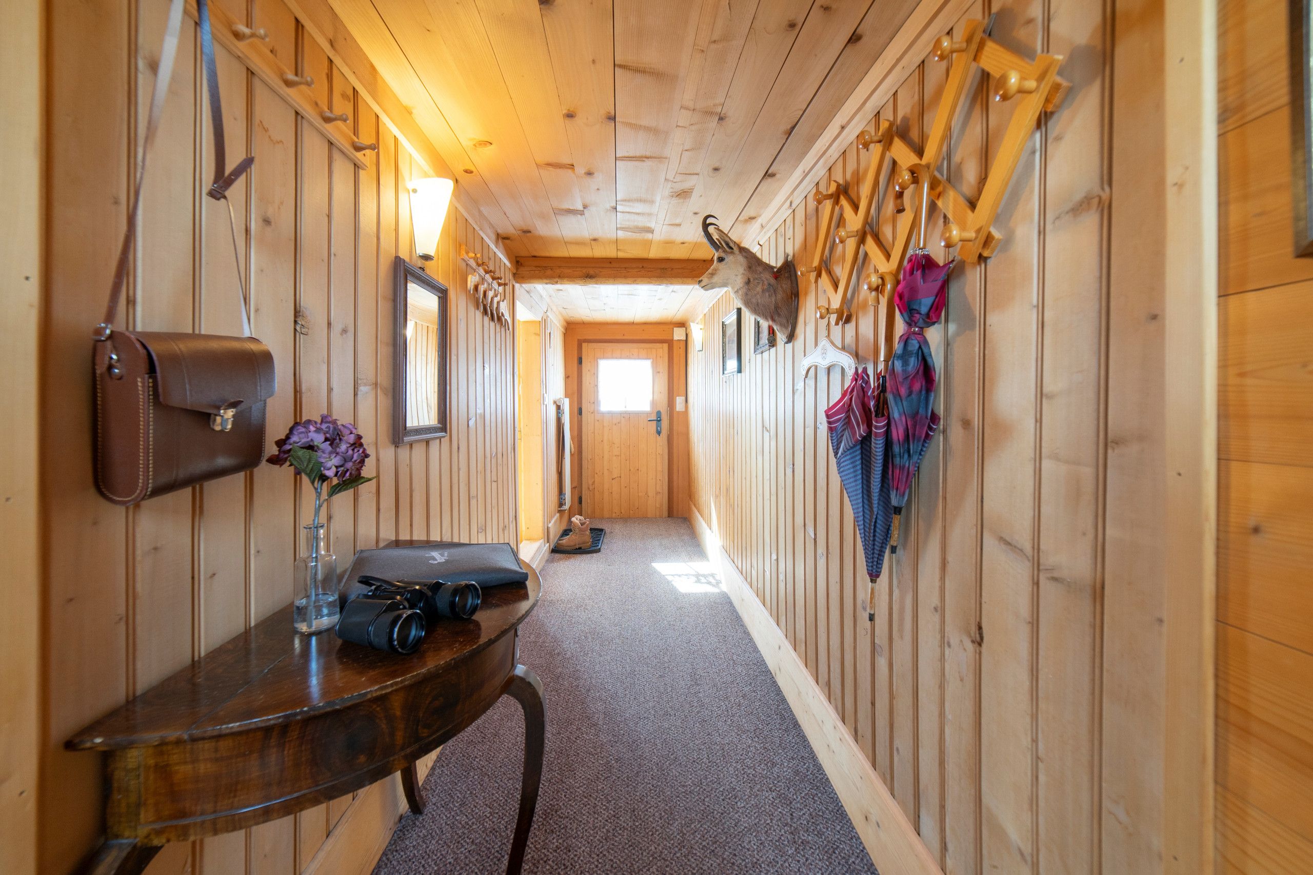 A wooden hallway with a table, a mirror, a mounted animal head, and a door at the end.