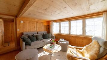 Ein gemütliches Zimmer mit Holzboden, einem Sofa, einem Sessel, einem kleinen Tisch und einem Fenster mit Vorhängen.