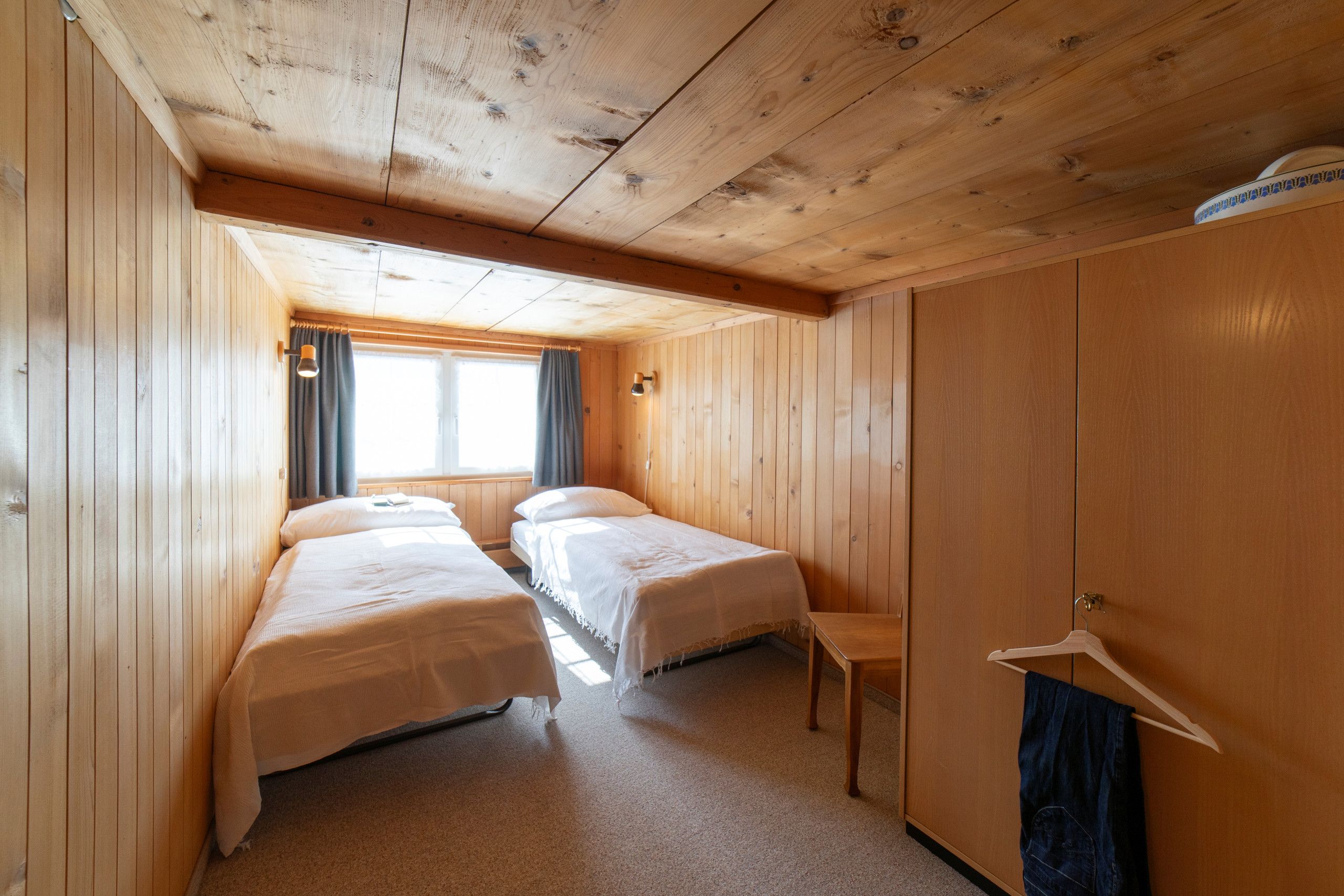 A room with two beds, a wooden wardrobe, a small table, and a window with curtains.