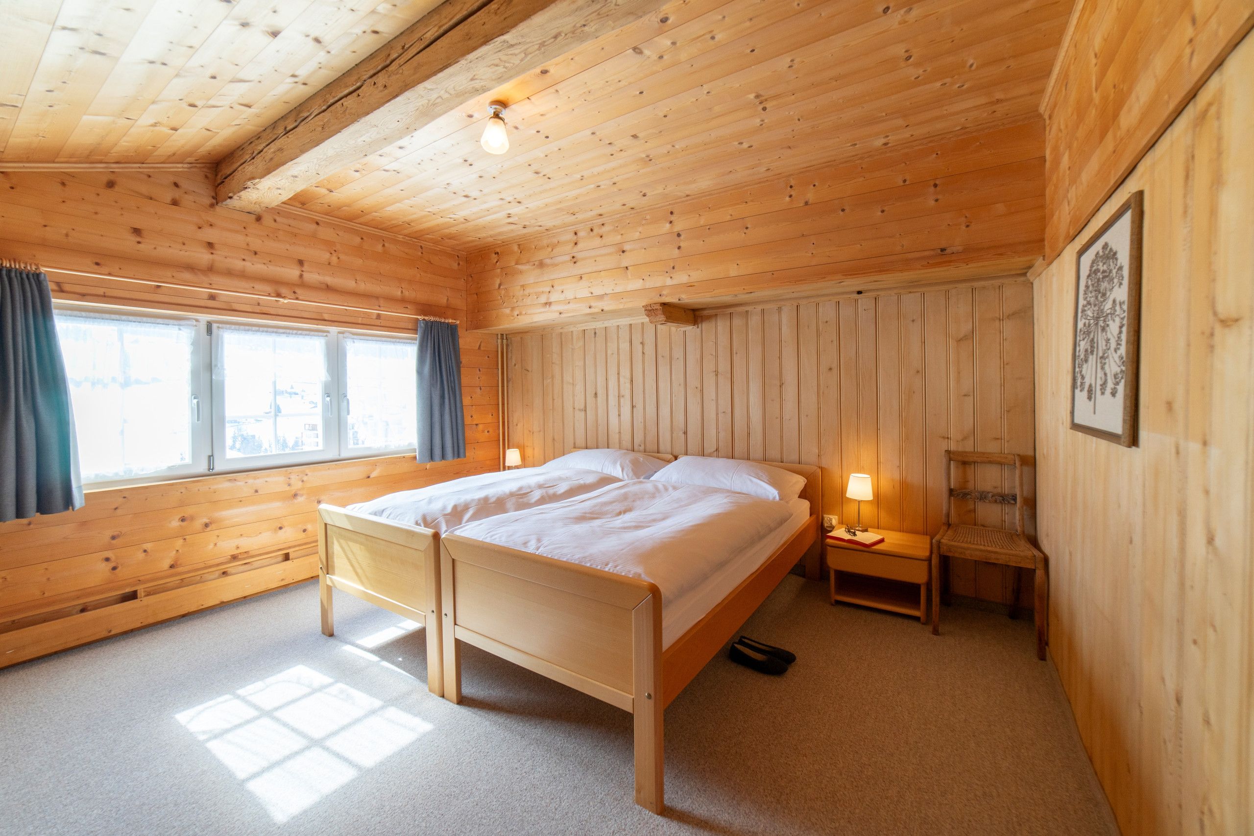 A wooden room with a bed, a chair, a nightstand, and a window.