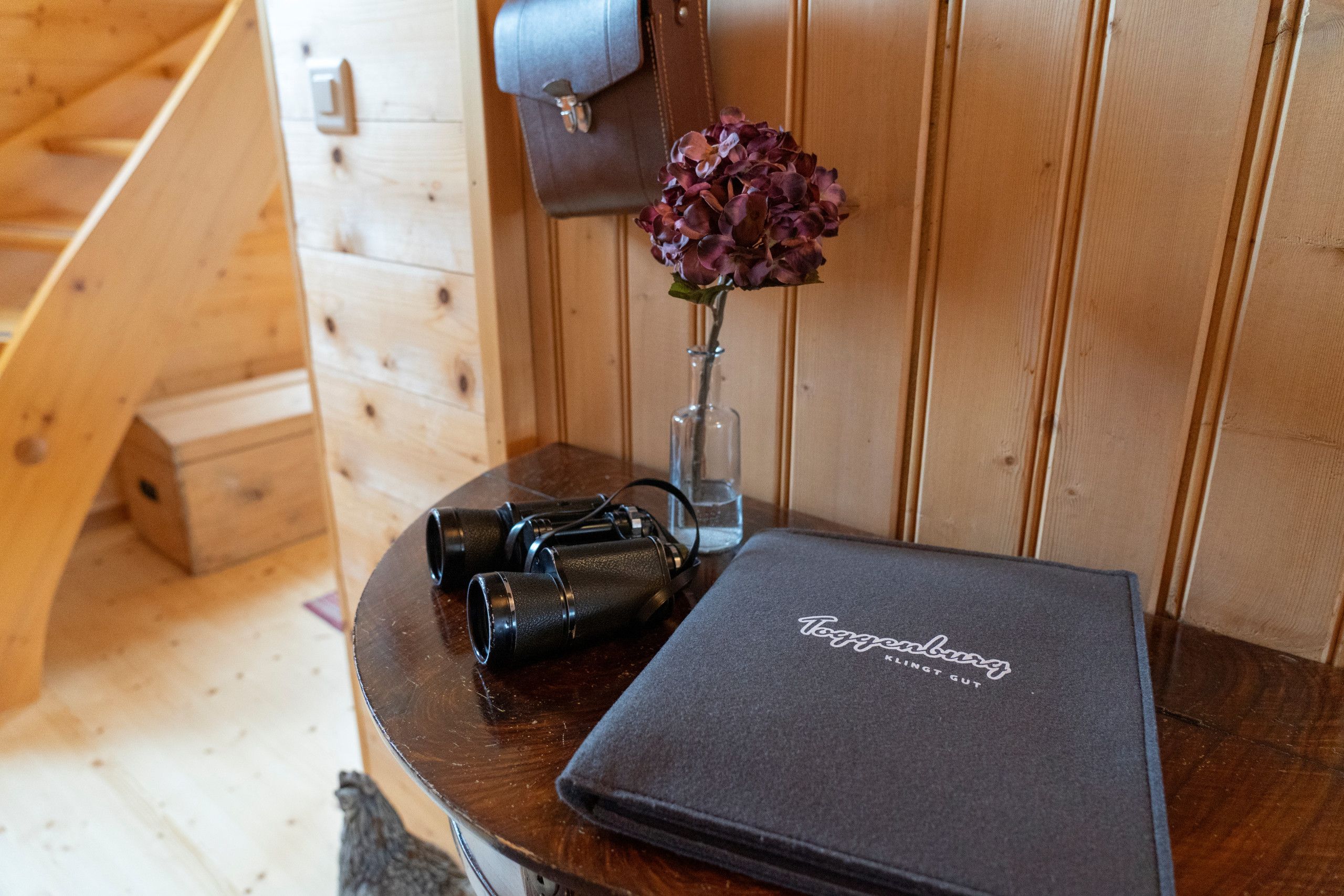 Ein Interieur einer Holzhütte mit einem Tisch, auf dem Ferngläser, eine Blumenvase und eine schwarze Tasche mit der Aufschrift 'Toyzeburg' liegen.