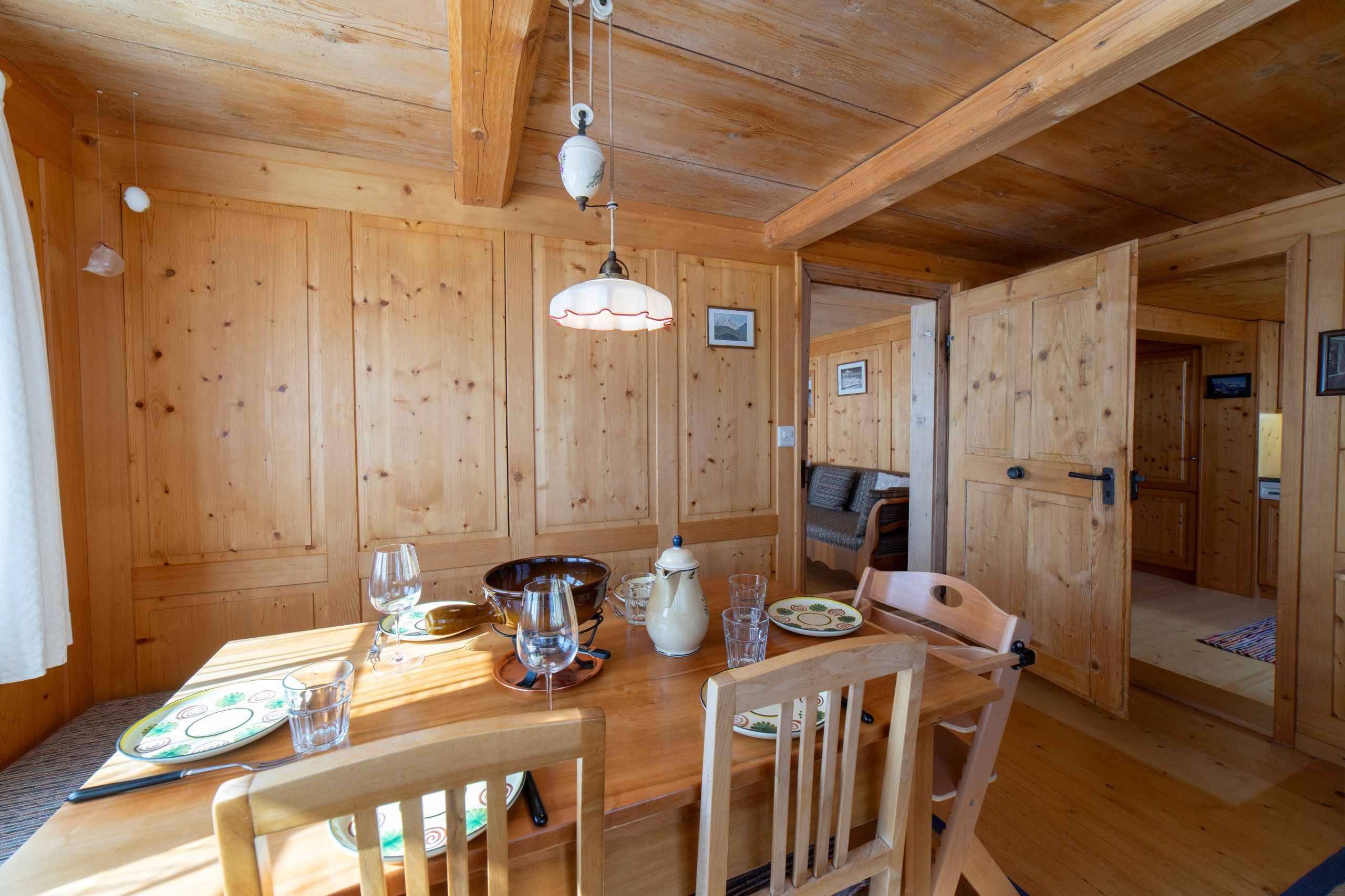 Un intérieur en bois avec une table à manger dressée pour deux personnes, une chaise, un luminaire suspendu et une porte donnant sur une autre pièce.