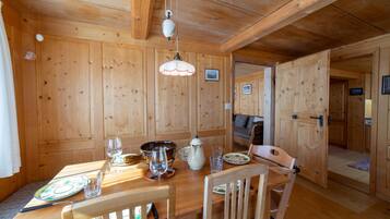 Un intérieur en bois avec une table à manger dressée pour deux personnes, une chaise, un luminaire suspendu et une porte donnant sur une autre pièce.
