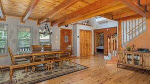 A spacious room with a wooden dining table, chairs, and a chandelier. There is a staircase with a wooden railing and a view of a garden through the windows. - Comfort Pines- Pinebrook Lake Access (Arnold)