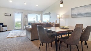 Uma sala de estar ampla com uma grande janela, uma área de jantar com mesa e cadeiras de madeira, e vista para o exterior.