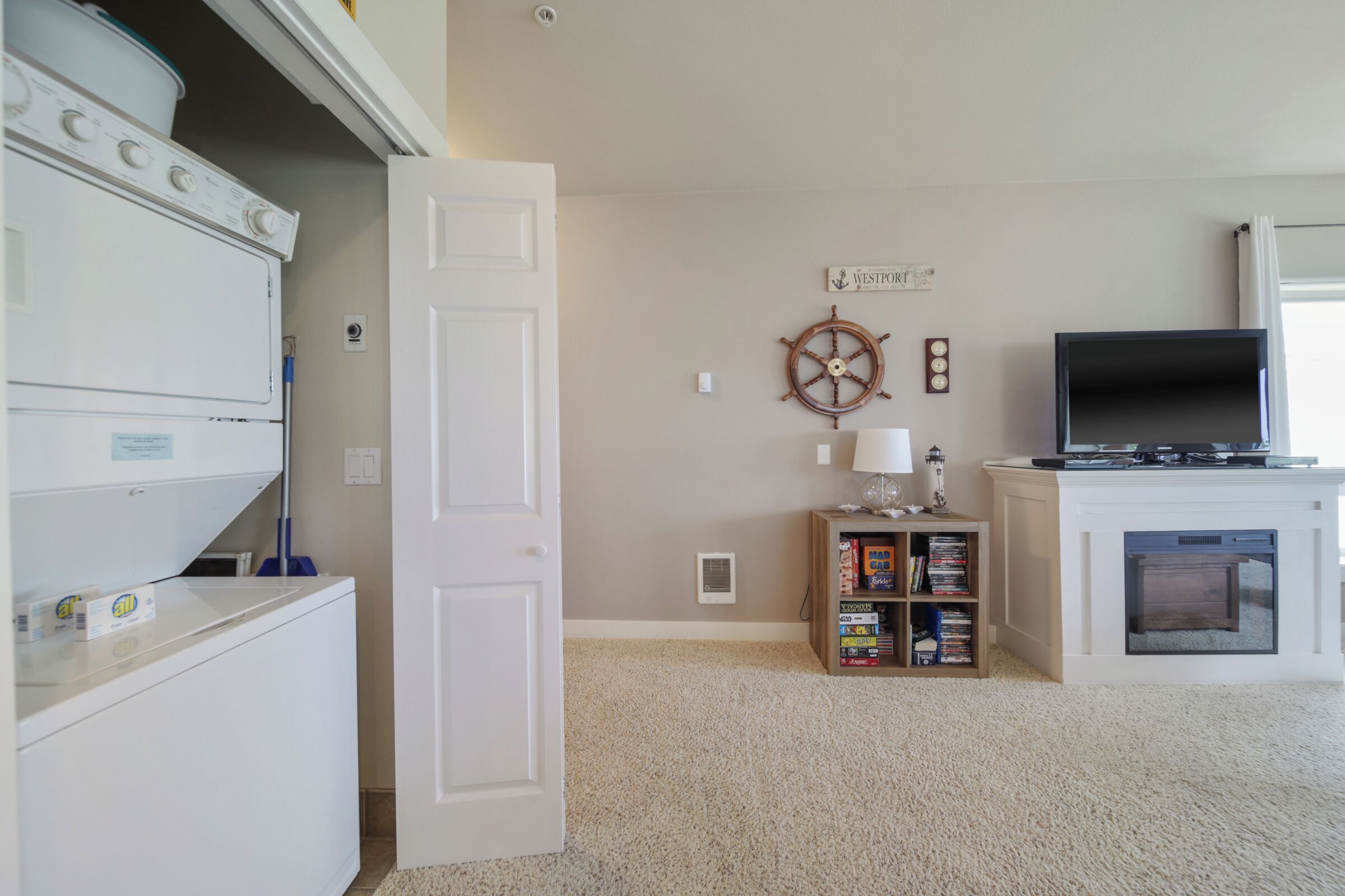A laundry area with stacked washers and dryers, a bookshelf with books and games, a wall-mounted ship's wheel, and a fireplace with a TV.