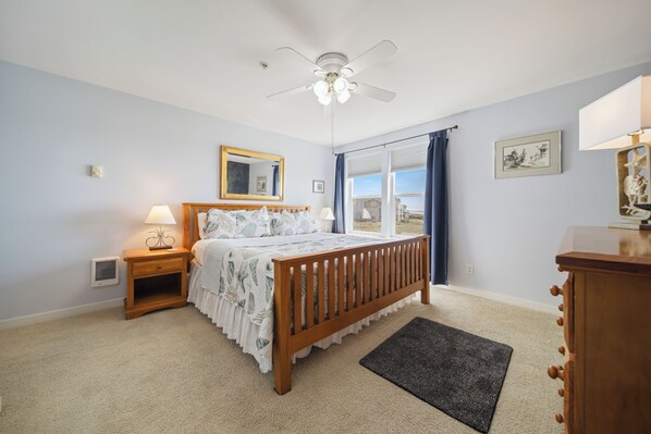 Ein Schlafzimmer mit Bett, Nachttisch, Kommode, Deckenventilator und großen Fenstern.