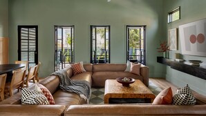 Ein modernes Wohnzimmer mit einem Ecksofa, einem Couchtisch und einem Essbereich mit einem Holztisch und Stühlen. Große Fenster sorgen für natürliches Licht und Ausblicke ins Freie.