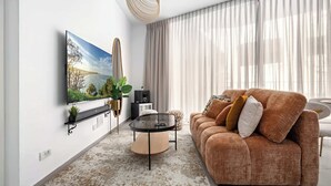 A living room with a sofa, a coffee table, a TV mounted on the wall, and a large window with curtains. - Comfortable apartment with Balcony by HolyGuest (Tel Aviv)