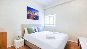 Hotellihuone, jossa on suuri sänky, yöpöytä lampun kanssa, ikkuna ja taulu seinällä.