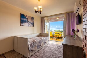 Ein kleines Zimmer mit Bett, Heizkörper, Fernseher, einem Fenster mit Vorhängen und einem Balkon mit Stuhl.