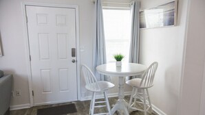 Una habitación pequeña y limpia con una puerta blanca, una mesa redonda con dos sillas blancas, una planta en maceta y una ventana con cortinas.