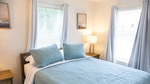 Un dormitorio con cama, dos almohadas, una mesita de noche, una lámpara y un cuadro en la pared.