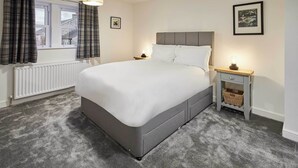 Una cama bien hecha en una habitación con cabecera gris, una mesita de noche con lámpara y una ventana con cortinas.